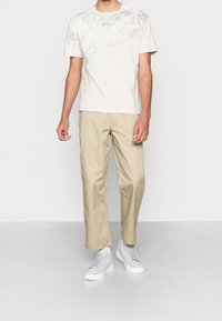 Krémové tričko s krátkým rukávem a texturovaným vzorem na hrudi, kombinované s béžovými kalhotami rovného střihu a bílými teniskami.