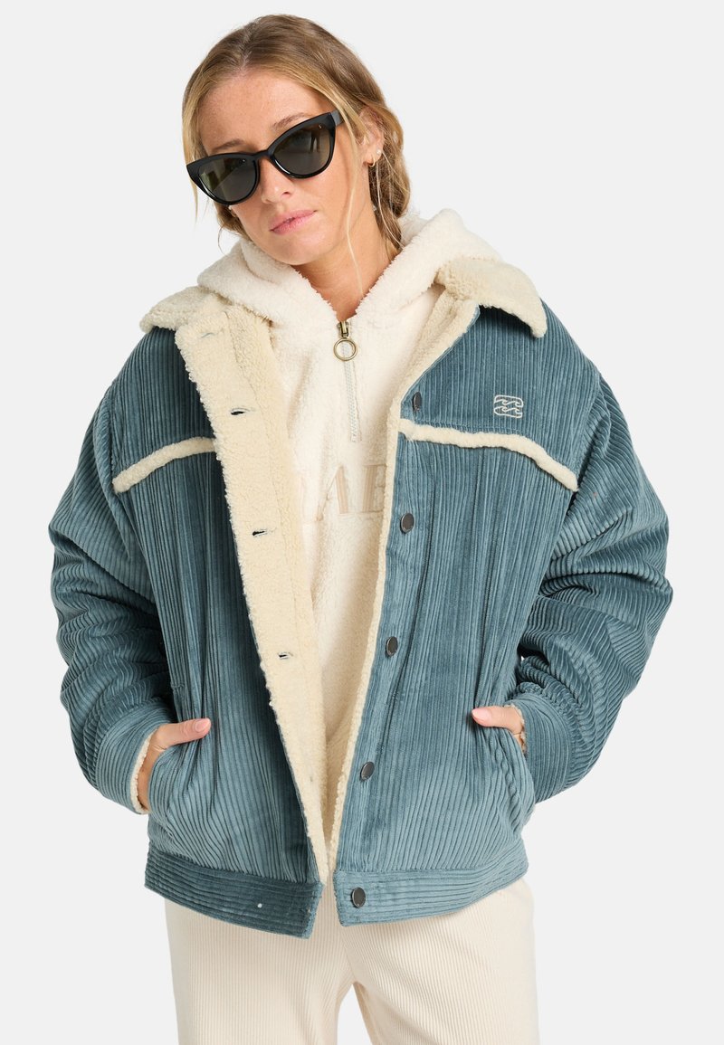 Overdimensioneret teal corduroy-jakke med beige sherpa-for og kontrasterende hvid syning, udstyret med tryklåse og sidelommer.