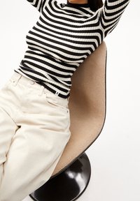 Haut à manches longues côtelé à rayures noires et blanches associé à un pantalon crème, assis sur une chaise ronde noire lisse avec un coussin beige texturé.