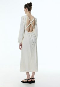 Langes, cremefarbenes Kleid mit langen Ärmeln, offener Rückseite und gekreuzten Trägern sowie einem strukturierten Stoff; kombiniert mit schwarzen Sandalen.