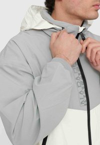 Giacca grigio chiaro con cappuccio bianco, chiusura con zip e dettagli neri. Caratterizzata da un tessuto testurizzato e un logo sul petto.
