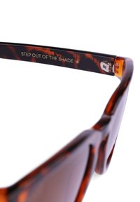 Sluneční brýle s želvovinovým vzorem, které mají silný rám, elegantní design a vyrytý text "VYSTUP Z OAZY." Obsahují kovové akcenty na závěsech.