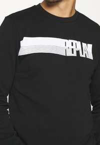 Camisa de manga larga negra con una franja horizontal blanca que presenta la palabra "PLAY" en letras negritas y líneas delgadas, hecha de una mezcla de algodón.