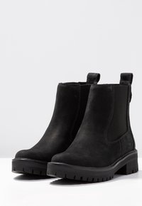 Mustat nahkaiset nilkkasaappaat, joissa on joustavat sivupaneelit, teksturoitu pinta ja kestävä kumipohja. Takana on vetolenksu.