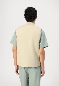 Beige, ujjatlan mellény magas gallérral, puha anyagból készült, világoszöld, rövid ujjú ing felett viselve. Egyszerű dizájn és laza fazon.