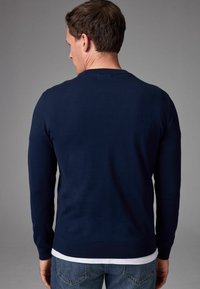 Maglione blu navy con maniche lunghe, collo giro, orlo a coste e texture liscia, indossato sopra una camicia bianca e abbinato a jeans blu.