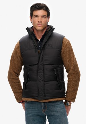 Black puffer vest with a zip front and two side pockets, paired with a brown fleece long-sleeve shirt. Features a high collar and quilted design.