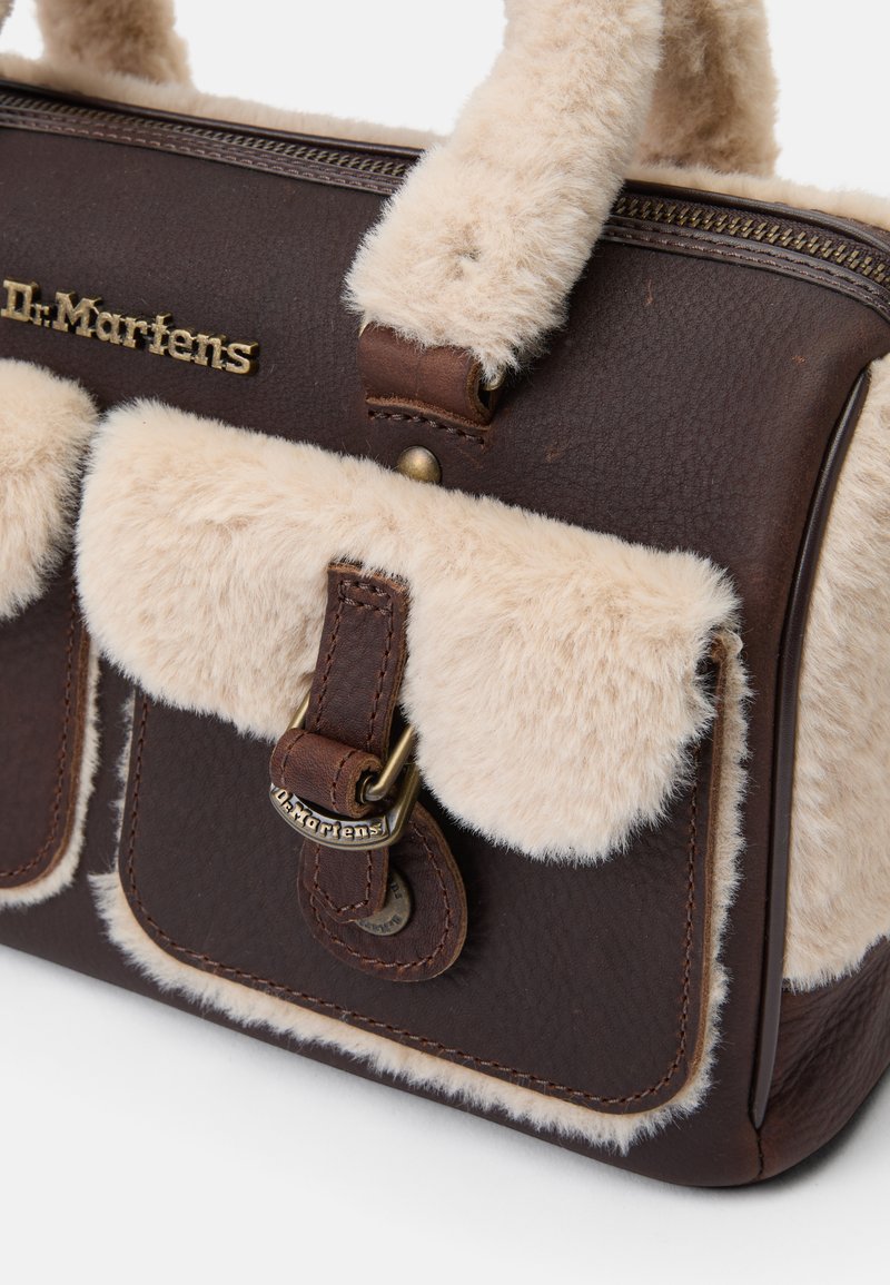 Bolso de mano de cuero marrón con acentos de pelaje sintético beige, que cuenta con dos bolsillos frontales, herrajes dorados y el logo de Dr. Martens en el lateral.