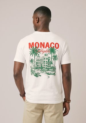 Muž nosiaci biele tričko s textom "MONACO Nights" a zelenou vintage ilustráciou budovy, paliem a klasického auta na chrbte.