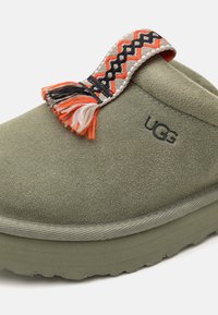 Zielono-oliwkowe zamszowe kapcie o teksturowanej powierzchni, z kolorowym plecionym paskiem akcentującym i frędzlami. Logo UGG widoczne.