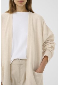 Cardigan bej din lână, cu mâneci lungi, deschis în față, cu două buzunare frontale, purtat peste un tricou alb și pantaloni de culoare bej deschis.