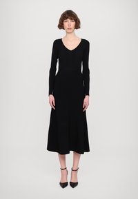 Abito nero a maniche lunghe con scollatura a V, corpino a costine e gonna fluida. Il tessuto appare liscio con una leggera elasticità. Abbinato a tacchi neri.