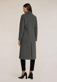 Grå uldfrakke med knapkrave, lange ærmer og sidelommer. Har en slids i ryggen for nemmere bevægelse. Bæres over sorte bukser og hæle.