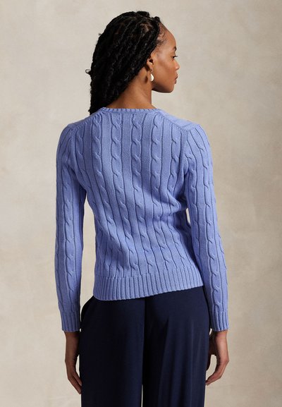 Kobieta z warkoczem widziana od tyłu, ubrana w jasnofioletowy sweter w warkocze i ciemne spodnie na neutralnym tle.