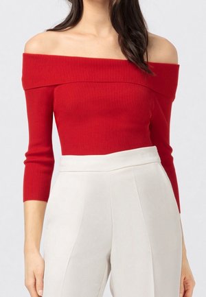 Femme portant un haut rouge côtelé à épaules dénudées avec des manches trois-quarts et un pantalon blanc taille haute sur un fond clair.