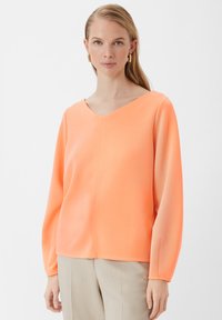 Lichtgewicht V-hals top in fel oranje met lange, wijdvallende mouwen, voorzien van een naadloos ontwerp en een zachte textuur. Gecombineerd met lichte broek.