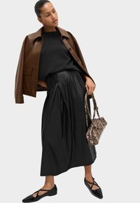 Veste en cuir marron, pull noir, jupe plissée noire, ballerines noires, portant un sac à main marron à motif serpent. Fond neutre.