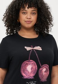 Svart t-skjorte med grafikk av to kirsebær med en rosa sløyfe, teksturert detailing, og setningen "La cerise sur le gâteau" under.