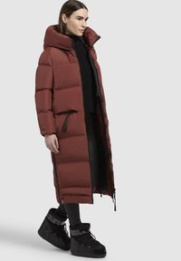 Lång vadderad kappa i burgundy med hög krage, dragkedja och sidofickor; matchad med svarta snörade vinterstövlar.