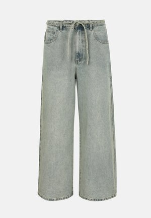 Magas derekú, bő szárú szürke farmernadrág elülső zsebekkel, övtartókkal és anyagból készült derékpánttal, egyszínű háttér előtt.