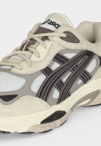 Μπεζ και γκρι παπούτσι τρεξίματος ASICS με mesh επάνω μέρος, σκουρόγκρι ρίγες, επενδεδυμένο γιακά και μαξιλαρωμένη σόλα.