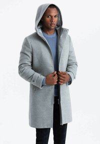 Abrigo gris con capucha y un diseño ajustado, que cuenta con un cierre de cremallera y botones a presión, combinado con un suéter azul de canalé por debajo.