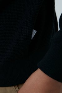 Granatowy sweter z teksturowanej tkaniny, z prążkowanym dołem i mankietami, z widoczną metką w biało-granatowy wzór.