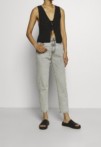 Black knitted vest with deep V-neck, cinched at the waist, paired with high-waisted light gray jeans and black slip-on sandals.