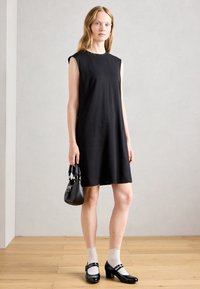 Femme se tenant à l'intérieur, portant une robe noire sans manches, des chaussettes blanches, des escarpins en cuir verni noir et tenant un petit sac à main noir.