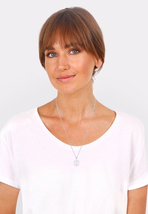 Silver necklace with an ornate round pendant on a thin chain, worn over a white scoop-neck t-shirt. Smooth finish with intricate details.