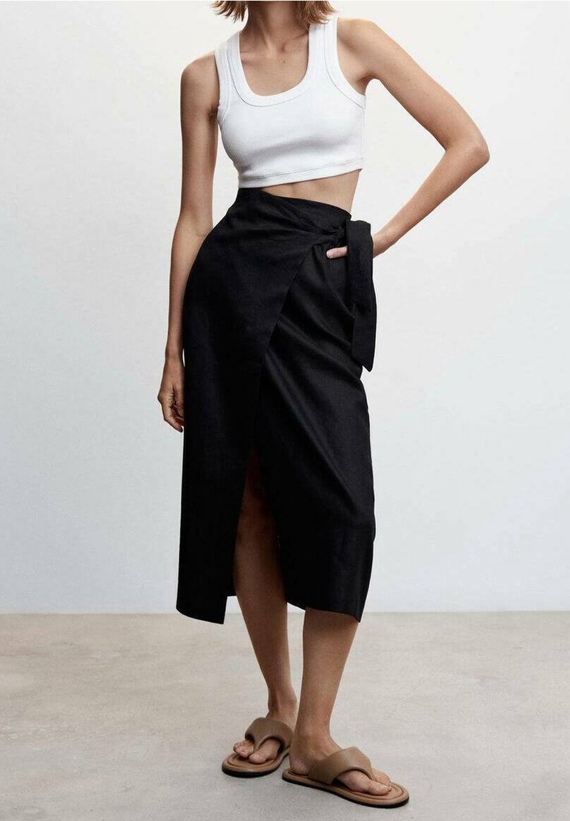 Femme portant un crop top blanc sans manches, une jupe portefeuille noire taille haute avec une fente, et des sandales marron à brides croisées, debout avec une main sur la hanche.