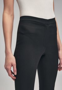 Pantalons noirs ajustés avec une texture lisse, taille haute et détails de couture sur les côtés, associés à un haut blanc côtelé visible au niveau de la taille.