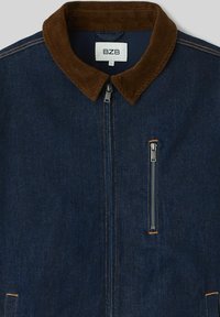 Veste en denim avec un col en velours côtelé marron, une fermeture éclair frontale et une poche poitrine zippée. Tissu bleu foncé avec une couture contrastante.