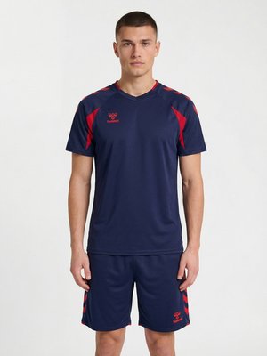 Ung mand står frontalt iført mørkeblå sportstøj med røde detaljer og Hummel-logoer på skjorte og shorts.