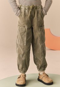 Helle olivgrüne Corduroy-Cargo-Hosen mit elastischen Bündchen und Taschen, kombiniert mit sandfarbenen Wildlederschuhen und cremfarbenen Socken auf einer kreisförmigen grünen Fläche.
