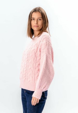 Młoda kobieta o prostych brązowych włosach, ubrana w jasnoróżowy, teksturowany sweter w warkocze oraz ciemnoniebieskie dżinsy, stojąca bokiem.