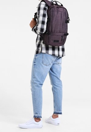 Zaino in tessuto texturizzato viola scuro, forma rettangolare, dotato di spallacci regolabili e molteplici tasche esterne. Indossato sopra una camicia a quadri.