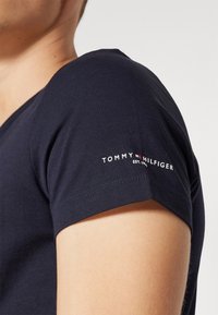 Námořnická modrá bavlněná trička s krátkým rukávem; má vyšitý logo "TOMMY HILFIGER" na rukávu v bílé a červené barvě.