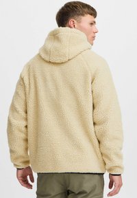 Beige fleece hettegenser med en teksturert, fluffy utside, med snor i hetten og elastiske mansjetter. Bakside viser en avslappet passform.