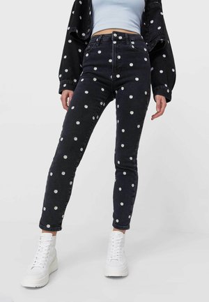 Person trägt hoch taillierte schwarze Jeans und Jacke mit weißen Gänseblümchen-Prints, hellblaues Croptop und weiße Plateau-Sneaker auf grauem Hintergrund.