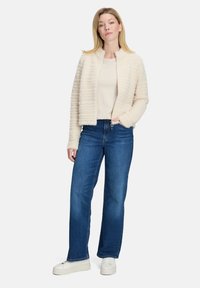 Puszysty kremowy kardigan z poziomymi teksturowanymi paskami, zestawiony z niebieskimi dżinsami z wysokim stanem i białymi sneakersami. Prosty okrągły dekolt.