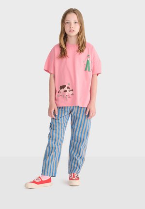 Ung pige iført en lyserød t-shirt med illustrationer af en bonde og en ko, blå og beige stribede bukser samt røde lærredssko med hvide såler.