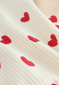 Kreedist kangas koorevärviga, millel on hajutatud punaste südamemustritega. Tekstuur tundub pehme ja kutsuv, väikese läikega.