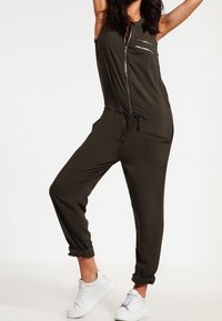 Olivgrüner Jumpsuit mit durchgehendem Reißverschluss, ärmellosem Design, verstellbarem Bund und umgeschlagenen Bündchen, kombiniert mit weißen Sneakern.