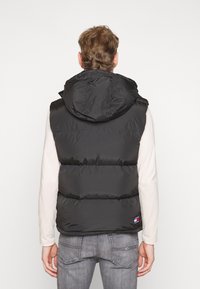 Schwarze Pufferweste mit Kapuze, gestepptem Design und sichtbaren Nähten. Beinhaltet ein Logo-Patch an der Taille, getragen über einem hellen Langarmshirt.