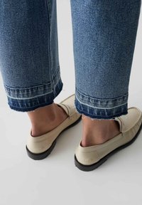 Scarpe beige slip-on con suole nere, abbinate a jeans blu in denim a vita alta con orlo sfrangiato e accenti in denim più chiaro.