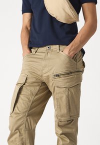 Beiga cargo byxor tillverkade av bomullsblandning, med flera fickor, dragkedjor och en avslappnad passform. Bärs med en marinblå t-shirt.