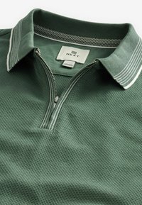 Vihreä polo-paita, joka on valmistettu teksturoidusta kankaasta ja jossa on puolivetoketju. Varustettu raidallisella kauluksella ja suoralla helmalla. Tunnuksessa lukee 'NEXT.'