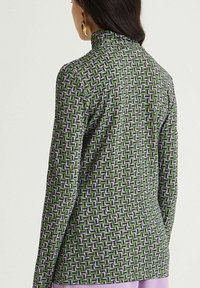 Haut à manches longues avec un motif géométrique vert et violet, doté d'un col montant et d'une texture de tissu lisse.