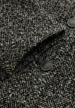 Wlněná bunda s texturovaným vzorem houndstooth v černé a béžové barvě, s černými kovovými knoflíky zdobenými složitými vzory.
